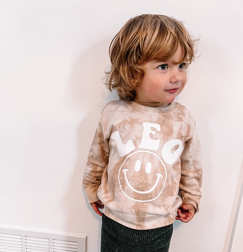 May include: A light brown tie-dye sweatshirt with a white smiley face and the name "LEO" in a retro font.
