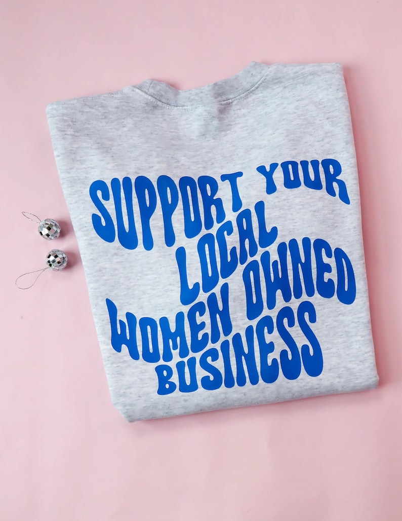 May include: A light gray sweatshirt with the blue text "SUPPORT YOUR LOCAL OWNED WOMEN BUSINESS". Two small silver disco ball ornaments are on the left side of the sweatshirt. The background is pink.
