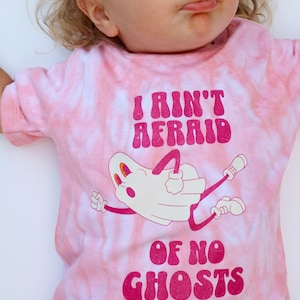 Puede incluir: Camiseta rosa te&ntilde;ida con la frase "I Ain't Afraid Of No Ghosts" en magenta. La camiseta presenta una ilustraci&oacute;n de fantasma de dibujos animados en blanco con detalles, brazos y piernas en magenta.