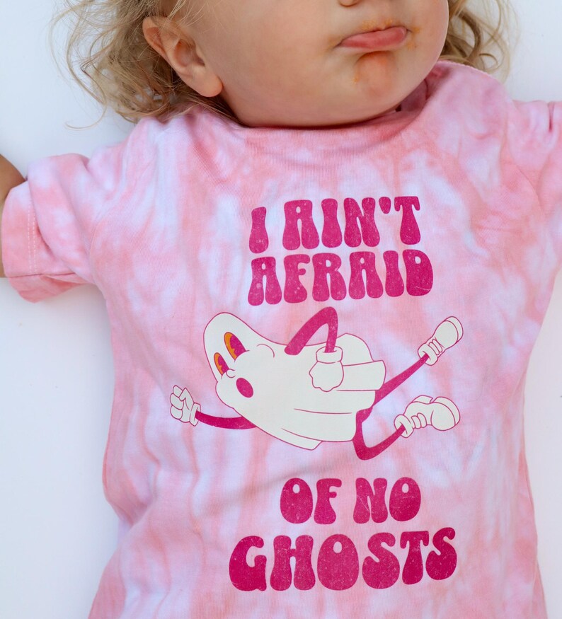 Puede incluir: Camiseta rosa te&ntilde;ida con la frase "I Ain't Afraid Of No Ghosts" en magenta. La camiseta presenta una ilustraci&oacute;n de fantasma de dibujos animados en blanco con detalles, brazos y piernas en magenta.
