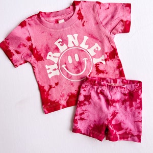 May include: Pink tie-dye short-sleeved shirt with a white smiley face and the text "RENLEY" in a retro font. Matching pink tie-dye shorts.
