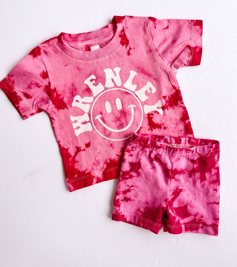 May include: Pink tie-dye short-sleeved shirt with a white smiley face and the text "RENLEY" in a retro font. Matching pink tie-dye shorts.