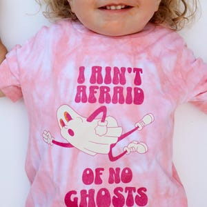 Puede incluir: Camiseta rosa te&ntilde;ida con la frase "I Ain't Afraid of No Ghosts" en letras rosa oscuro. Una ilustraci&oacute;n de fantasma de dibujos animados aparece debajo del texto. La camiseta tiene un patr&oacute;n de remolino rosa claro y blanco.