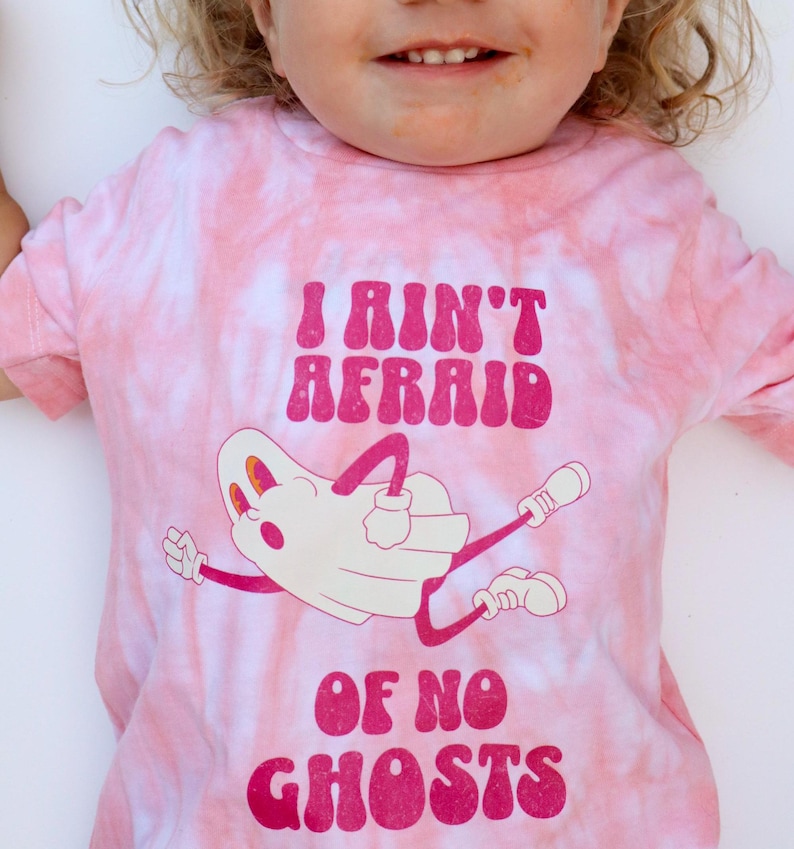 Puede incluir: Camiseta rosa te&ntilde;ida con la frase "I Ain't Afraid of No Ghosts" en letras rosa oscuro. Una ilustraci&oacute;n de fantasma de dibujos animados aparece debajo del texto. La camiseta tiene un patr&oacute;n de remolino rosa claro y blanco.