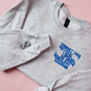 May include: A heather gray sweatshirt with the text "SUPPORT YOUR LOCAL WOMEN OWNED BUSINESS" in blue. The sweatshirt has long sleeves and a crew neck. Two disco ball earrings are on the sleeve. The background is pink.