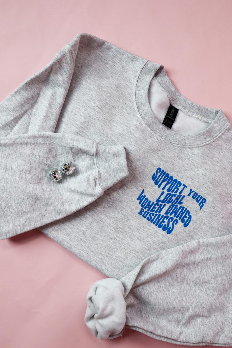May include: A heather gray sweatshirt with the text "SUPPORT YOUR LOCAL WOMEN OWNED BUSINESS" in blue. The sweatshirt has long sleeves and a crew neck. Two disco ball earrings are on the sleeve. The background is pink.