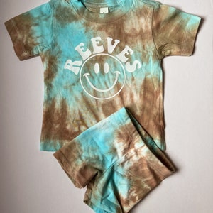 May include: A brown and blue tie-dye t-shirt with short sleeves and a white smiley face with the word "REEVES" above it. The t-shirt is paired with matching brown and blue tie-dye shorts.