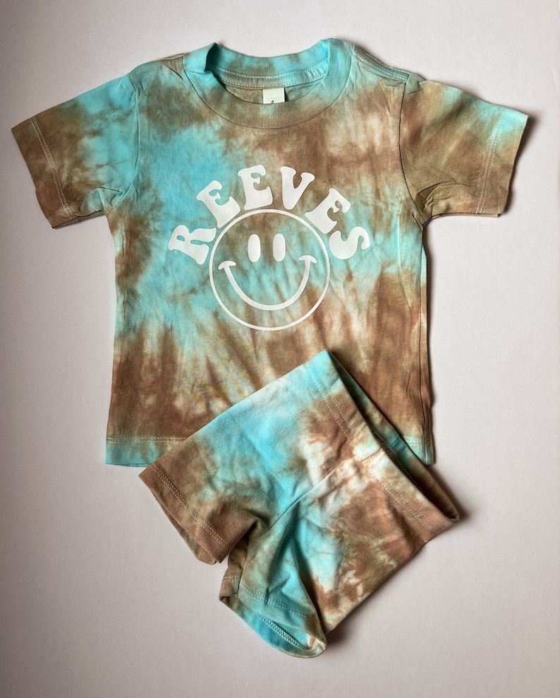 May include: A brown and blue tie-dye t-shirt with short sleeves and a white smiley face with the word "REEVES" above it. The t-shirt is paired with matching brown and blue tie-dye shorts.