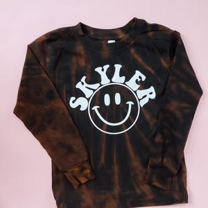 May include: A black and brown tie-dye long-sleeve shirt with a white smiley face and the name "Skyler" in a retro font.