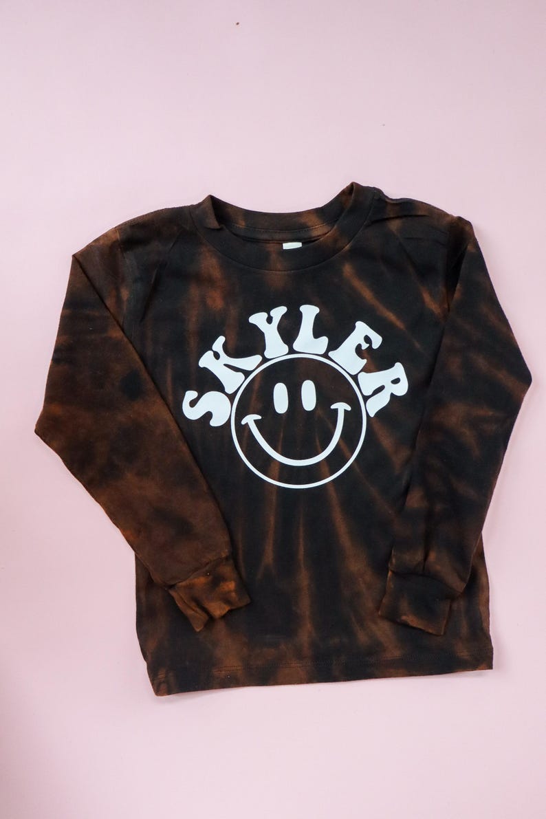 May include: A black and brown tie-dye long-sleeve shirt with a white smiley face and the name "Skyler" in a retro font.