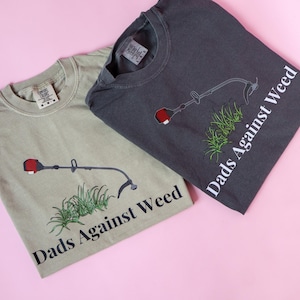 Pu&ograve; includere: Due t-shirt con una grafica di un decespugliatore e il testo "Dads Against Weed". Una maglietta &egrave; verde chiaro e l'altra &egrave; grigio scuro.