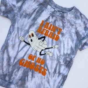 Puede incluir: Una camiseta te&ntilde;ida con tinte gris y blanco con un fantasma de dibujos animados y el texto naranja "I Ain't Afraid Of No Ghosts". El fantasma tiene una expresi&oacute;n de sorpresa y est&aacute; en movimiento.
