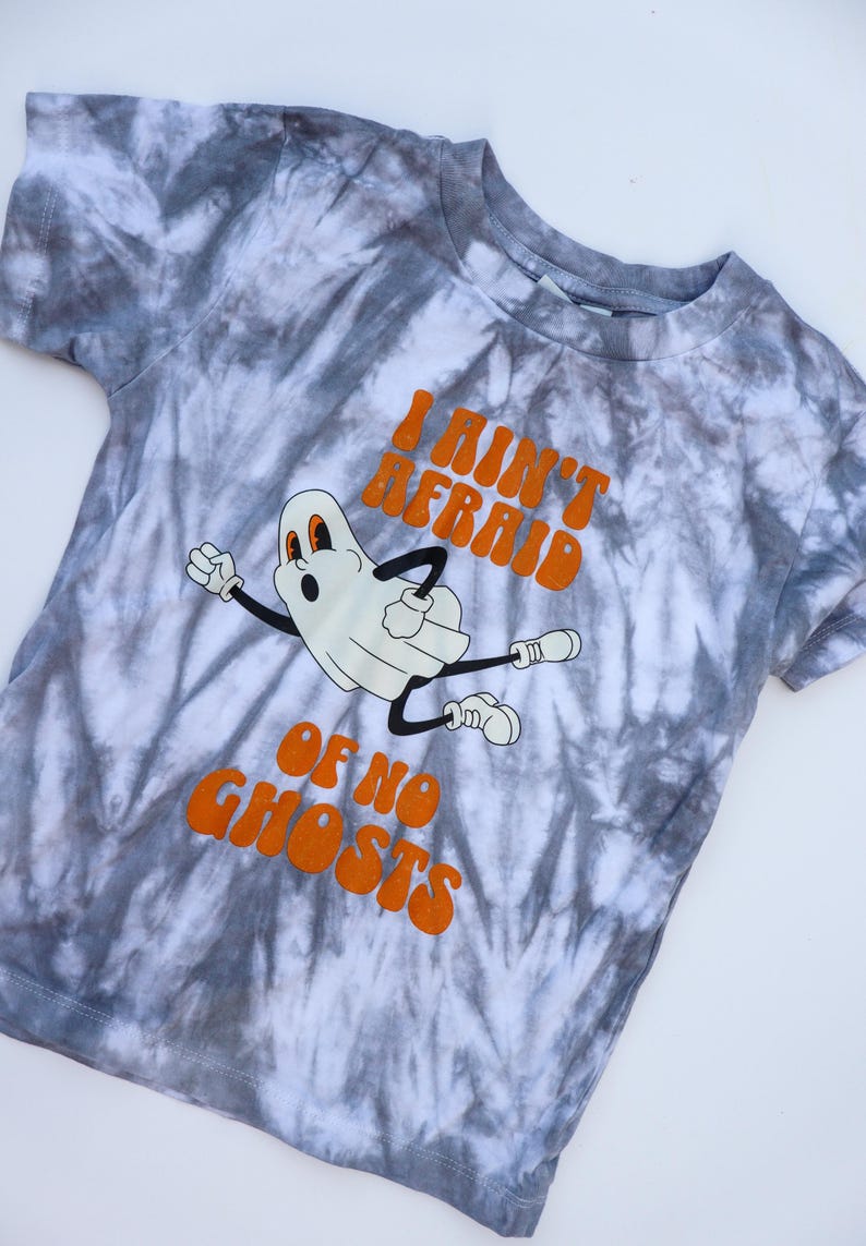 Puede incluir: Una camiseta te&ntilde;ida con tinte gris y blanco con un fantasma de dibujos animados y el texto naranja "I Ain't Afraid Of No Ghosts". El fantasma tiene una expresi&oacute;n de sorpresa y est&aacute; en movimiento.