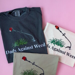 Pu&ograve; includere: Tre magliette con una grafica di un decespugliatore e il testo "Dads Against Weed". Le magliette sono di colore grigio antracite, beige e bianco.