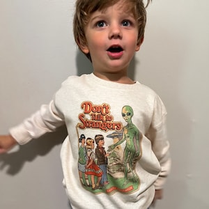 以下が含まれることがあります： クリーム色のスウェットシャツを着た幼い子供が、宇宙人と子供たちのグラフィックプリントを着ています。グラフィックには「見知らぬ人と話さないでください」と「安全教育」と書かれています。
