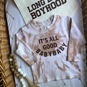 Può includere: Una maglietta a maniche lunghe con stampa tie-dye marrone e bianca con il testo "It's all good baby baby" stampato in marrone. La maglietta è appoggiata su uno sfondo bianco con un tessuto marrone e bianco con il testo "Long Live Boyhood" stampato in marrone.
