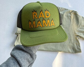 Pode incluir: Um boné de caminhoneiro verde com uma malha preta nas costas. O boné tem as palavras "RAD MAMA" em letras laranja com chamas nas letras.