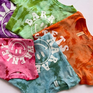 May include: Five tie-dye t-shirts with different colors and designs. Each shirt features a white smiley face with text around it. The text on the shirts reads "BARR", "HAPPY", "STAY", "LETS", and "FAR".
