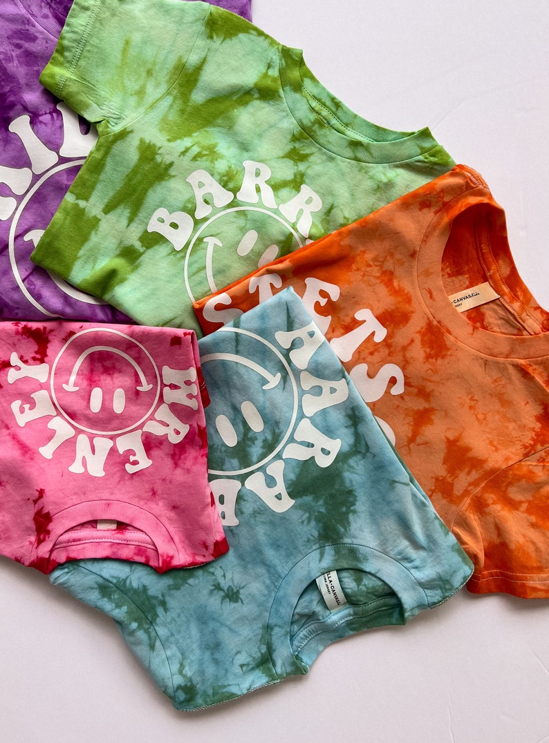 May include: Five tie-dye t-shirts with different colors and designs. Each shirt features a white smiley face with text around it. The text on the shirts reads "BARR", "HAPPY", "STAY", "LETS", and "FAR".