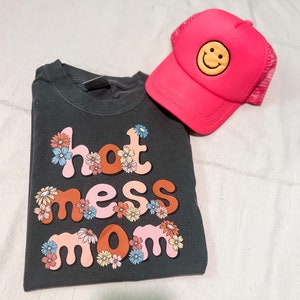 Pode incluir: Uma camiseta cinza escura com o texto "hot mess mom" em letras coloridas com flores ao redor das letras. Também há um boné de caminhoneiro rosa com um rosto sorridente amarelo na imagem.
