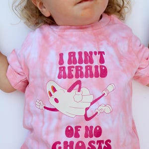 Puede incluir: Camiseta rosa te&ntilde;ida con la frase "I Ain't Afraid Of No Ghosts" en magenta. La camiseta presenta una ilustraci&oacute;n de fantasma de dibujos animados en blanco y magenta. La camiseta est&aacute; dise&ntilde;ada para ni&ntilde;os.