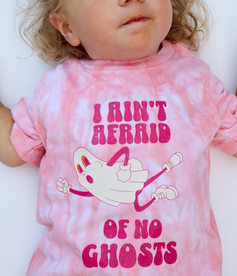 Puede incluir: Camiseta rosa te&ntilde;ida con la frase "I Ain't Afraid Of No Ghosts" en magenta. La camiseta presenta una ilustraci&oacute;n de fantasma de dibujos animados en blanco y magenta. La camiseta est&aacute; dise&ntilde;ada para ni&ntilde;os.