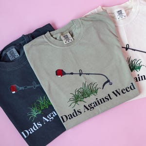 Pu&ograve; includere: Tre magliette con un'immagine di un decespugliatore e il testo "Dads Against Weed". Le magliette sono di colore nero, verde e bianco.
