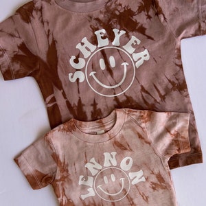 May include: Two brown tie-dye t-shirts with white smiley faces and text. The larger shirt says "SCHEYER" and the smaller shirt says "WENNON".