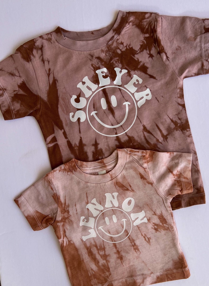 May include: Two brown tie-dye t-shirts with white smiley faces and text. The larger shirt says "SCHEYER" and the smaller shirt says "WENNON".
