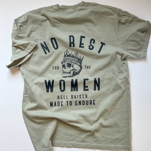 May include: A green t-shirt with black text that reads "No Rest For The Women, Hell Raiser Made To Endure". A skull with a crown is above the text.