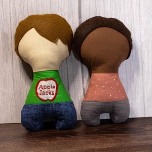 May include: Two handmade fabric dolls with brown and black hair. One doll wears a green shirt with "Apple Jacks" and blue denim pants. The other doll has a peach-colored dotted shirt and gray pants. The dolls are on a wooden surface.
