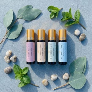 Puede incluir: Cuatro botellas de aceite esencial en roll-on con tapas doradas, etiquetadas como "Muscle Relief", "Alleviate Headache", "Digestion Aid" y "Breathe Easy". Las botellas están rodeadas de hojas de eucalipto, ramitas de menta y pequeñas piedras. Cada botella contiene 10 ml de aceite esencial.