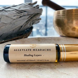 Könnte beinhalten: Eine Glas-Rollerflasche für ätherisches Öl mit goldenem Verschluss und einem Etikett mit der Aufschrift "ALLEVIATE HEADACHE" und "Healing Layers". Die Flasche enthält 10 ml ätherisches Öl. Im Hintergrund ein Salbeibündel und eine Klangschale aus Messing.