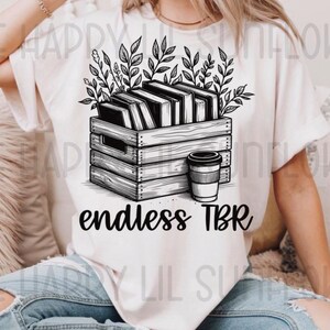 Könnte beinhalten: Ein weißes T-Shirt mit einem schwarzen Grafikdruck einer Holzkiste voller Bücher und Blattzweige, begleitet von einem Kaffeebecher. Der Text "endless TBR" ist in Schwarz unter der Kistenillustration auf der Vorderseite des Shirts gedruckt.