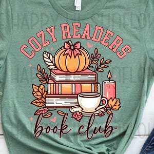 May include: A sage green t-shirt featuring a graphic print of a stack of books, a pumpkin with a pink bow, a cup of coffee, and a candle. The text reads COZY READERS book club in pink and black lettering. The design includes autumn leaves.