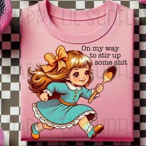 Könnte beinhalten: Rosa T-Shirt mit einer Cartoon-Illustration eines Mädchens, das mit einem Löffel rennt, der Text lautet "On my way to stir up some shit." Das T-Shirt ist von Discokugeln umgeben.