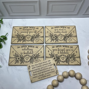 Puede incluir: Cuatro placas rectangulares de madera grabadas con diseños de sobres y el texto Open When, seguido de frases sobre la vida adulta, los choques de realidad y la valentía. Una etiqueta de madera más pequeña con texto está en primer plano.