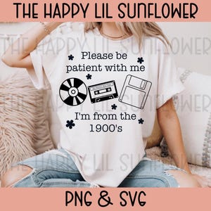 May include: A white short-sleeve t-shirt featuring black text that reads "Please be patient with me I'm from the 1900's" along with black graphic illustrations of a compact disc, a cassette tape, a floppy disk, and small flower icons.