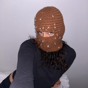 May include: Brown crocheted balaclava with clear crystal embellishments. The head covering is made of a textured, knitted material. The balaclava covers the head and neck, with openings for the eyes and mouth.