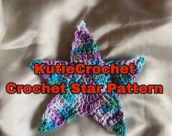 May include: A crocheted star in shades of purple, blue, green, and white. The star is on a white fabric background. The text "KutieCrochet Crochet Star Pattern" is displayed below the star.
