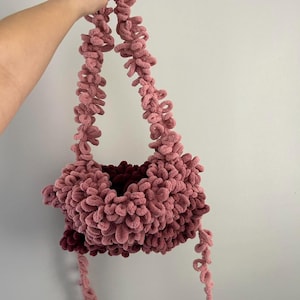 May include: A handmade, fluffy pink shoulder bag with a unique texture. The bag features a looped design on the straps and body, with a darker pink interior. The bag is held up by a hand.
