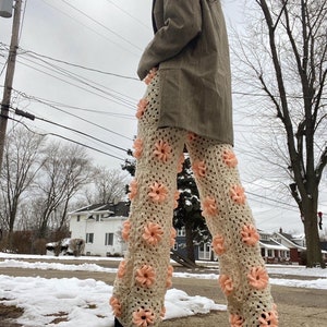 May include: A pair of crocheted pants with a floral design. The pants are cream colored with pink flowers. The person is wearing a brown blazer.