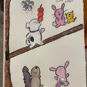 May include: Hand-drawn illustration on cream-colored paper featuring a dog balancing red fruit, a pink bunny, a yellow cat, and a squirrel on a brown line. A silver flower and a pearl are attached to the paper. The text "<clap clap>" is above the bunny and cat.