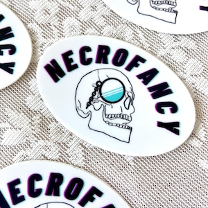 May include: White oval sticker with a black and white skull wearing a monocle with a blue lens. The text "NECROFANCY" is written in black with a purple outline around the letters.