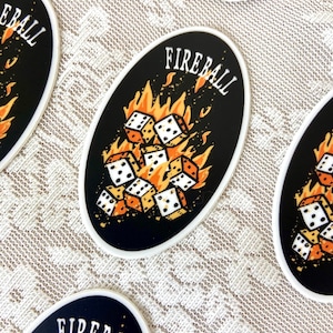 Op de afbeelding: Een ovale sticker in zwart en wit met de tekst "FIREBALL" in witte letters. De sticker toont een stapel witte dobbelstenen met zwarte stippen, die door oranje vlammen worden verzwolgen.