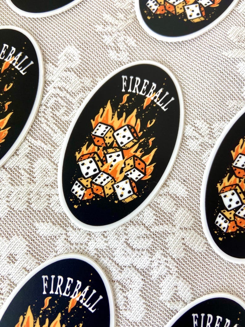 Op de afbeelding: Een ovale sticker in zwart en wit met de tekst "FIREBALL" in witte letters. De sticker toont een stapel witte dobbelstenen met zwarte stippen, die door oranje vlammen worden verzwolgen.