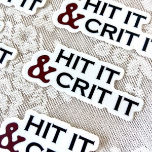 以下が含まれることがあります： 「& HIT IT CRIT IT」と黒文字で書かれた白いステッカー。アンパサンドはマルーンです。