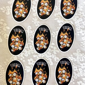 Op de afbeelding: Negen ovale stickers met een zwarte achtergrond en witte dobbelstenen in brand. De tekst "FIREBALL" staat in oranje bovenaan elke sticker.
