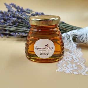 May include: A small, clear glass jar filled with golden honey, topped with a gold lid. The jar has a label that reads "Lavender & Honey Creek Farm, Ltd." and "Net Wt. 3 oz." A bundle of lavender and lace are in the background.
