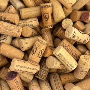 May include: A close-up shot of a pile of used wine corks. The corks are various sizes and shapes, with visible text and branding from different vineyards. The colors are primarily shades of brown and beige, with some showing purple staining.
